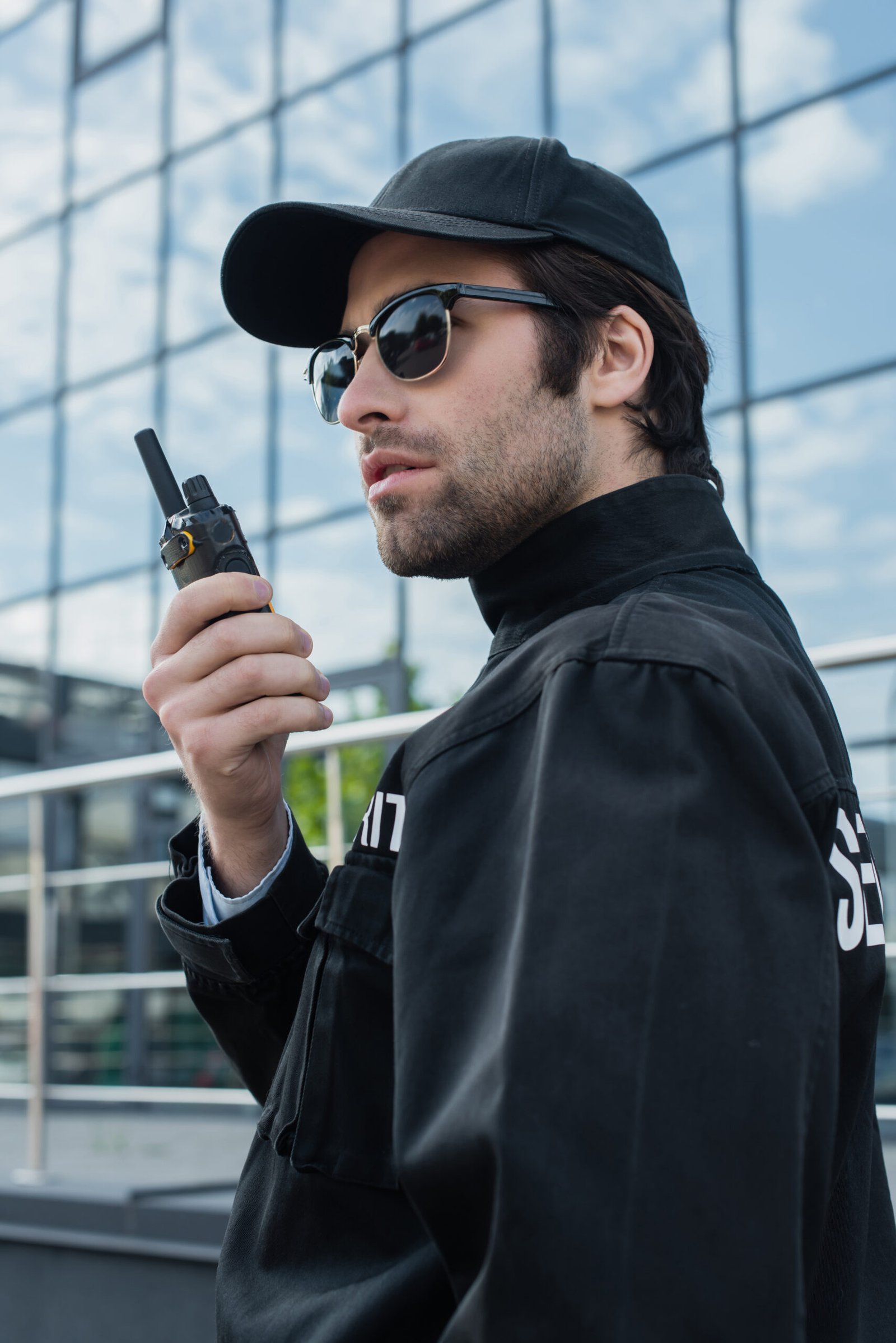 serious guard in sunglasses and black uniform talking on radio set outdoors serious guard in sunglasses and black uniform talking on radio set outdoors